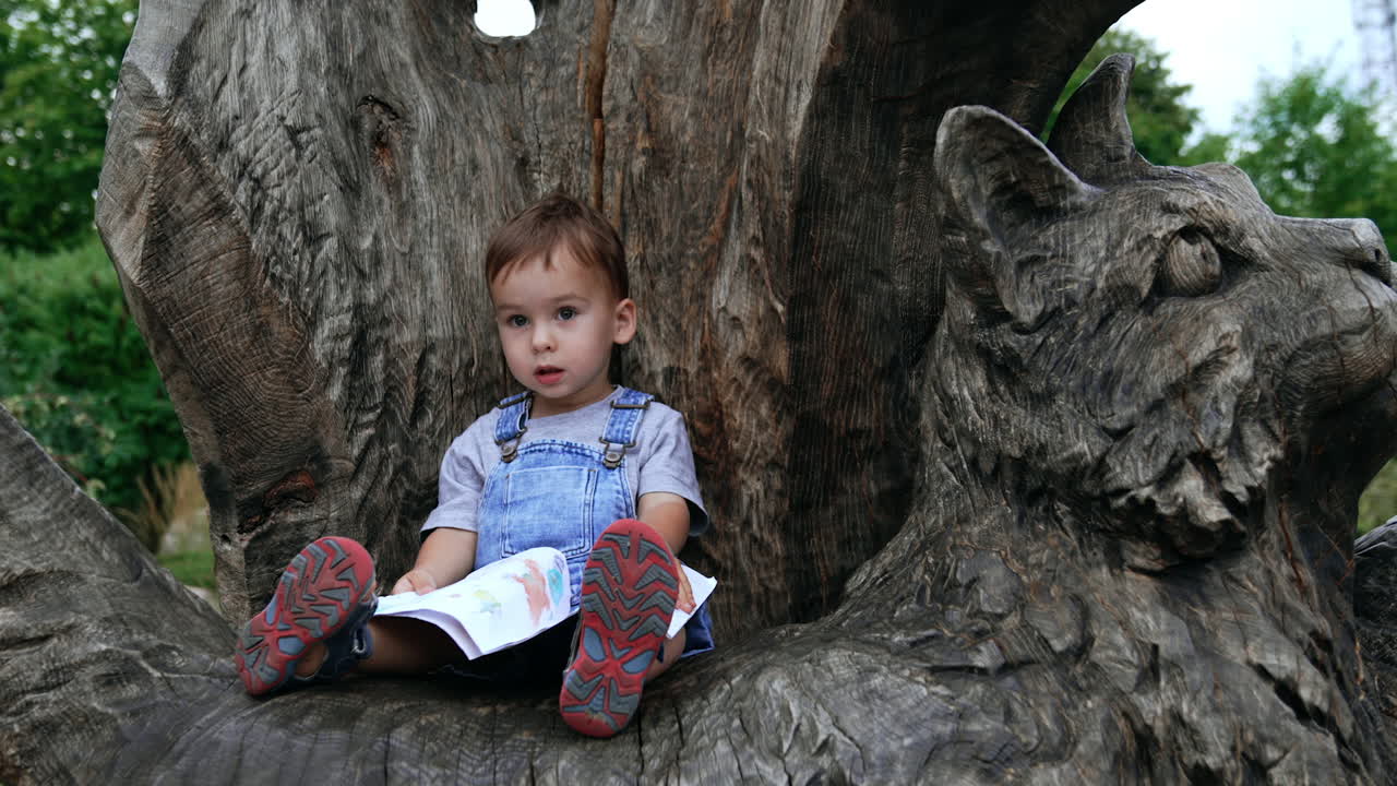 Lovely baby boy in jeans romper sitting on the wooden sculpture of a big cat. Active kid waves with a book he's holding in his hands.