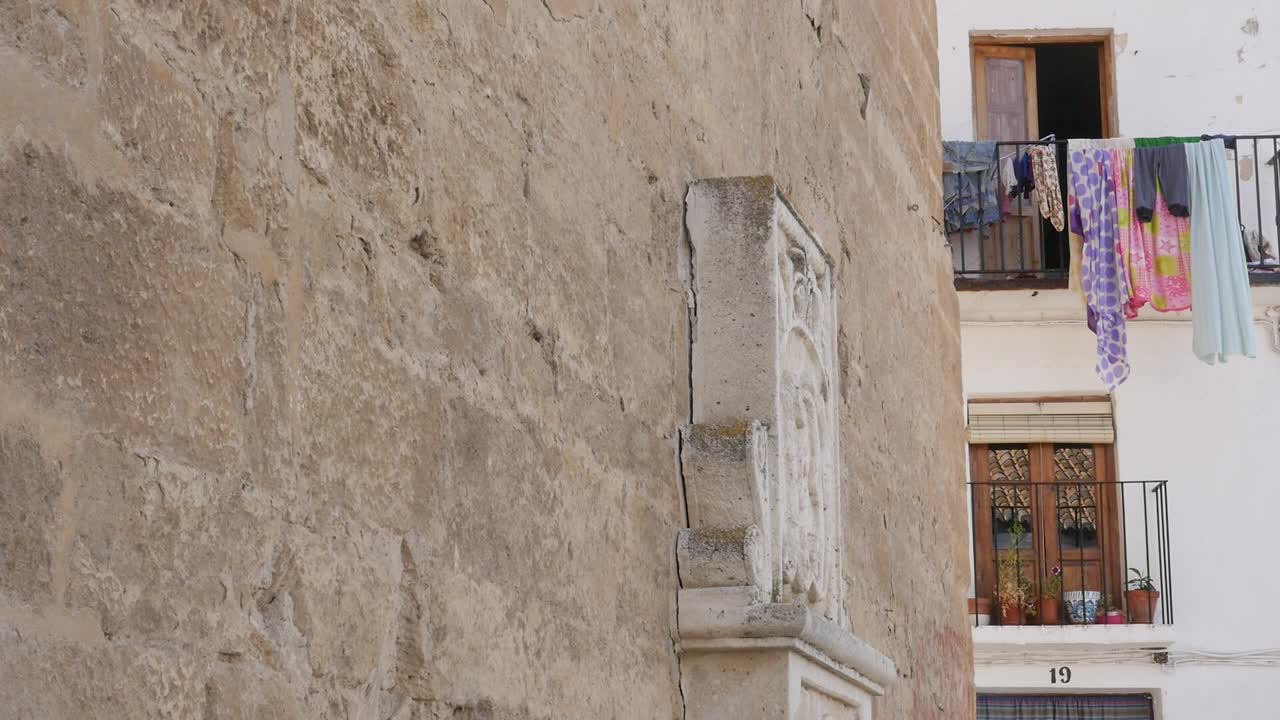 Primo piano di un muro di pietra storico con un rilievo religioso accanto a una casa tradizionale con panni stesi su un balcone. Luce naturale, inquadratura statica. Concetto di tradizione e vita quotidiana.
