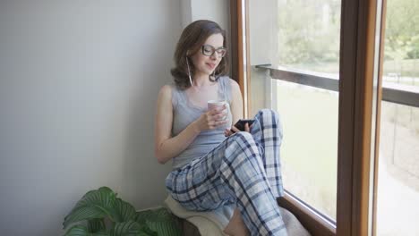smiling woman sitting in window using smartphone and earphones