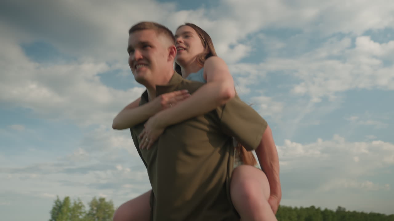 man carrying woman piggyback across green field under bright sky, sunlit clouds backdrop, joyful affectionate moment captured in playful outdoor exercise