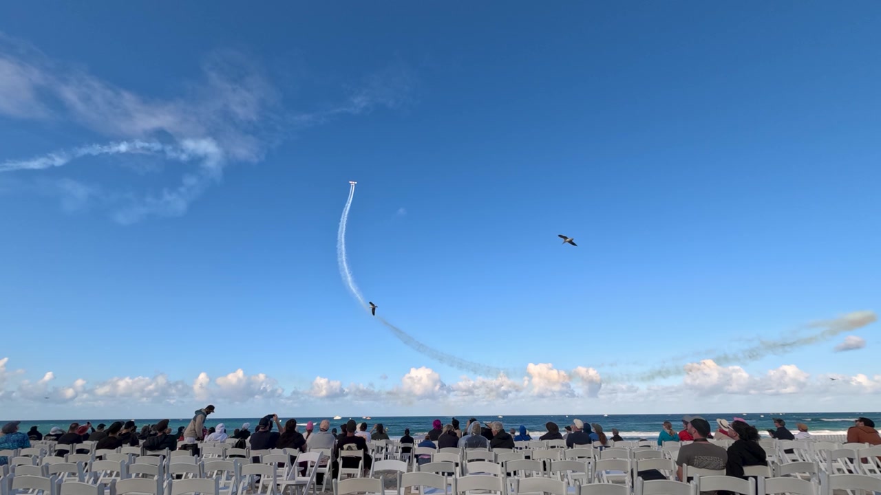 Spectators seated on Surfers Paradise beach watch a stunt plane leave smoke trails during airshow.