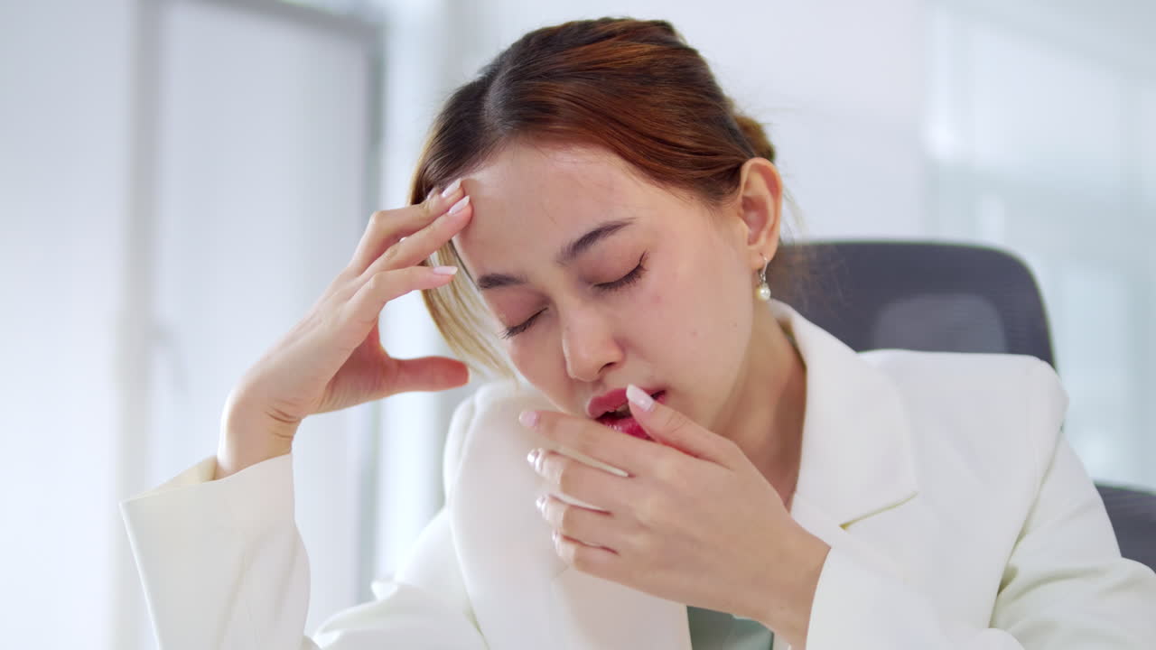 Holding her forehead as she tries to fight her sleepiness, a female office worker looks tired and sleepy.
