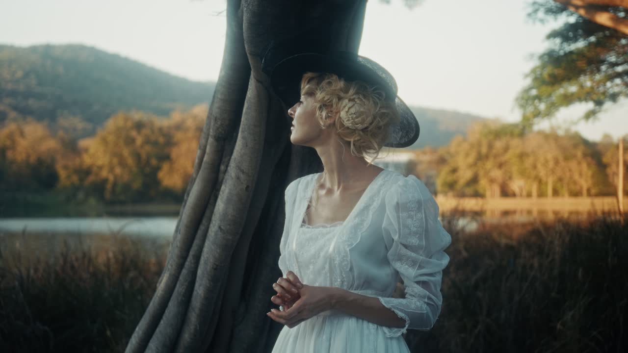 Elegantní žena ve vintage šatech a klobouku stojící u jezera při západu slunce