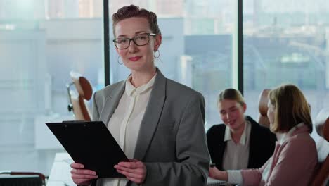 Retrato-De-Una-Chica-Rubia-De-Mediana-Edad-Segura-De-Sí-Misma-Con-Gafas-Y-Ropa-De-Trabajo-Sosteniendo-Una-Tableta-En-Sus-Manos-Cerca-De-Sus-Colegas-Empresarias-En-Una-Oficina-Moderna-Con-Grandes-Ventanales