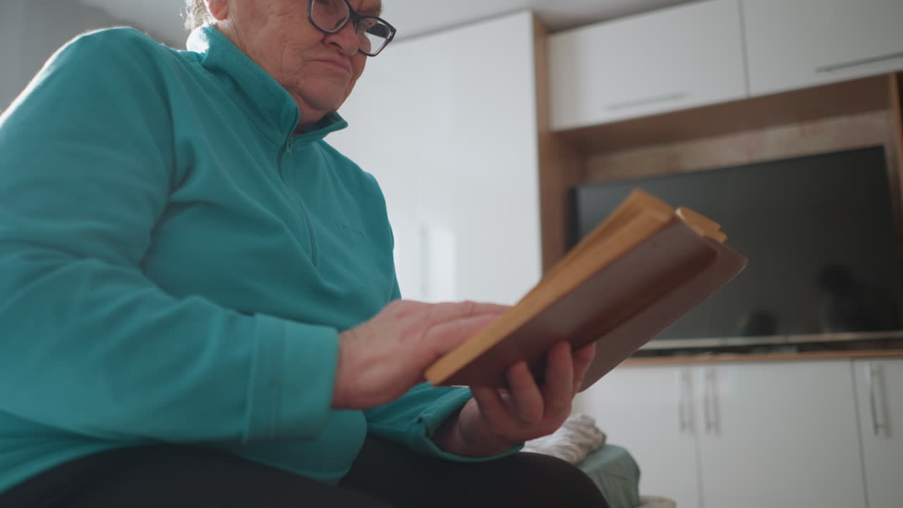Senior woman in teal sweater sitting on bed, focused on reading a hardcover book. Sunlight floods the room, highlighting her concentration in a peaceful and cozy atmosphere