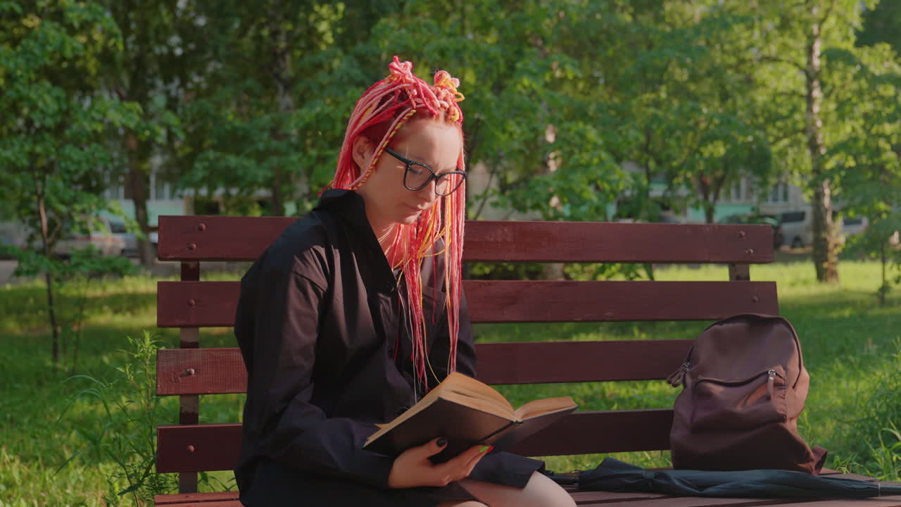 joven disfrutando de un libro en un entorno natural, mujer absorta en la lectura de un libro sentada en un banco rodeada de vegetación, joven sentada en un banco del parque absorta en su libro bajo la luz moteada del sol
