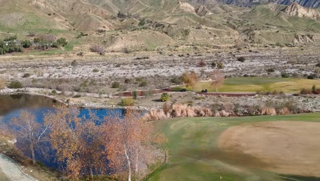 Eine-Drohne-Erhebt-Sich-Hinter-Einem-Busch-Und-Zeigt-Berge-Und-Drei-Golfwagen,-Die-über-Einen-Pfad-Auf-Einem-Wunderschönen-Golfplatz-Rasen