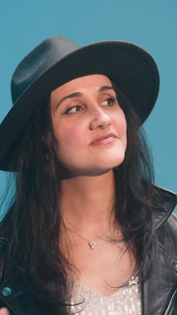 dama con una chaqueta de cuero negra y sombrero negro sonriendo mientras ajusta su cabello. en el fondo, un tecladista está borroso contra un telón de fondo azul, añadiendo profundidad a la escena