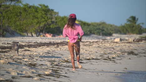 fit latin girl in a bikini and a shirt running on the beach with her dog pet enjoying and having fun