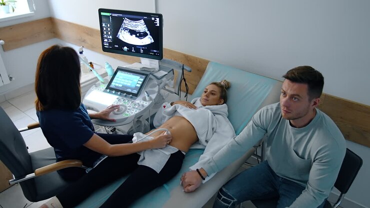 Future parents are in the doctor's cabinet at pregnancy check up. Caucasian lady lies on the couch undergoing ultrasound examination. Worried man holding wife's hand. Top view.