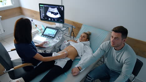 future parents are in the doctor's cabinet at pregnancy check up. caucasian lady lies on the couch undergoing ultrasound examination. worried man holding wife's hand. top view.
