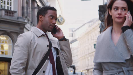 young black businessman standing on street talking on smartphone, white millennial woman walking past also using phone, close up, low angle