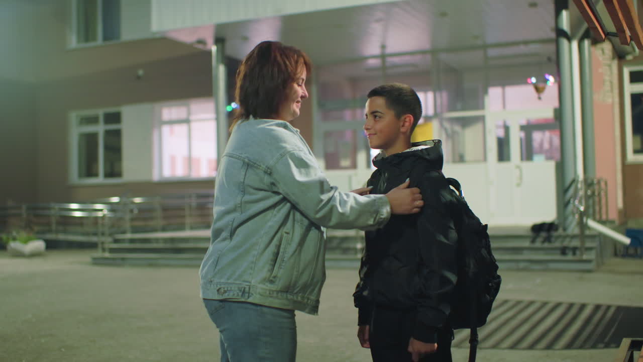 Woman kneels in front of boy outside school entrance early morning, adjusting his jacket with care as he prepares for day, soft lighting and empty courtyard highlight quiet emotional connection