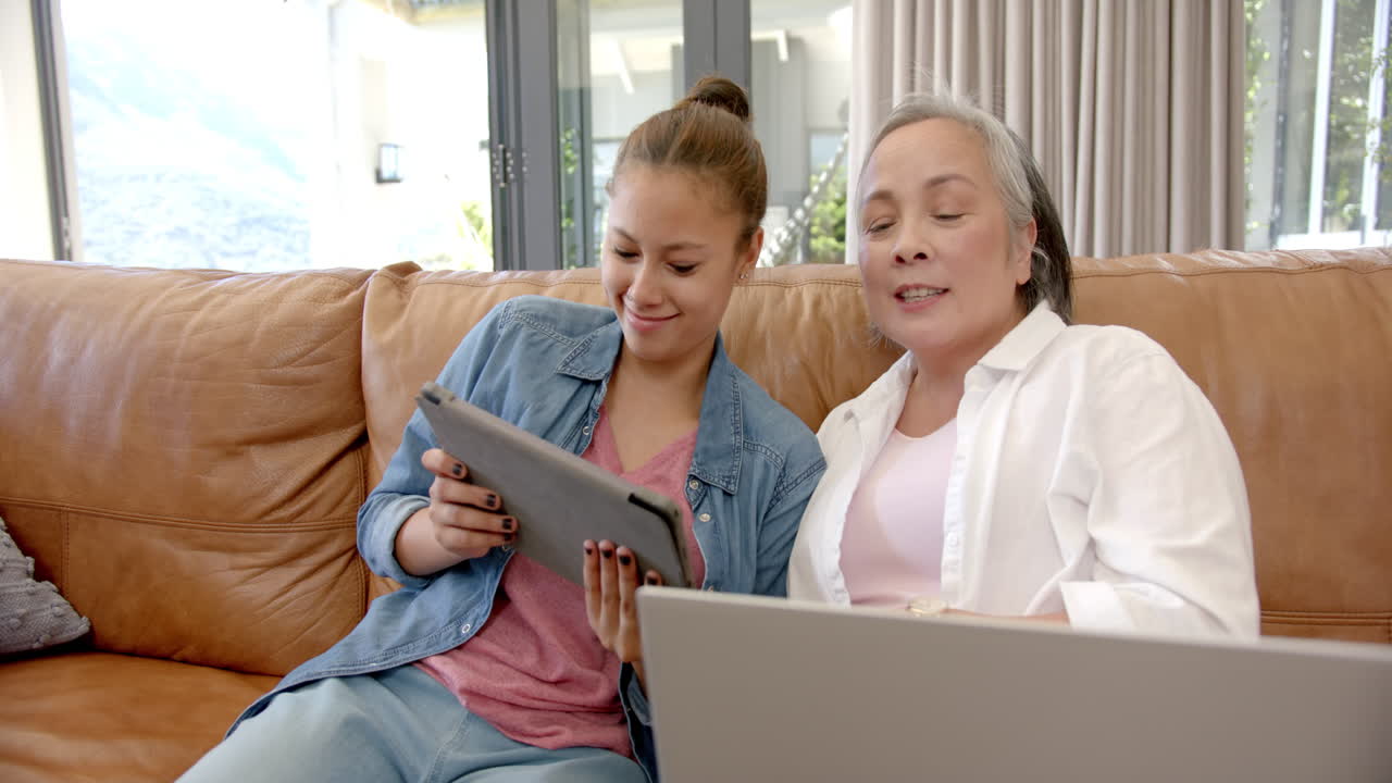 Using tablet and laptop, asian grandmother and granddaughter bonding on couch at home