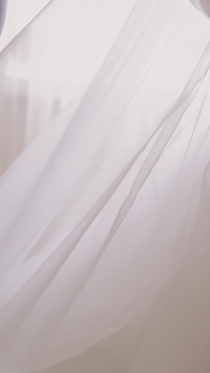 CU, slow motion: mother in light blue suit adjusts long transparent veil of young daughter wearing beautiful white lacy wedding dress at home closeup
