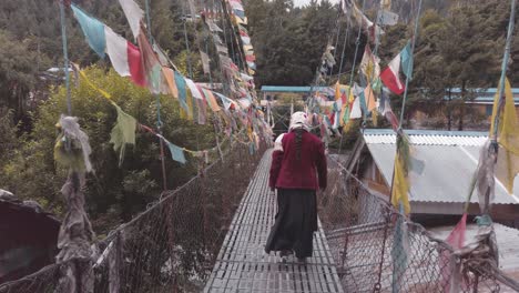 mujer nepal tradicional cruzando un puente colgante de hierro con bandera de colores en medio del himalaya