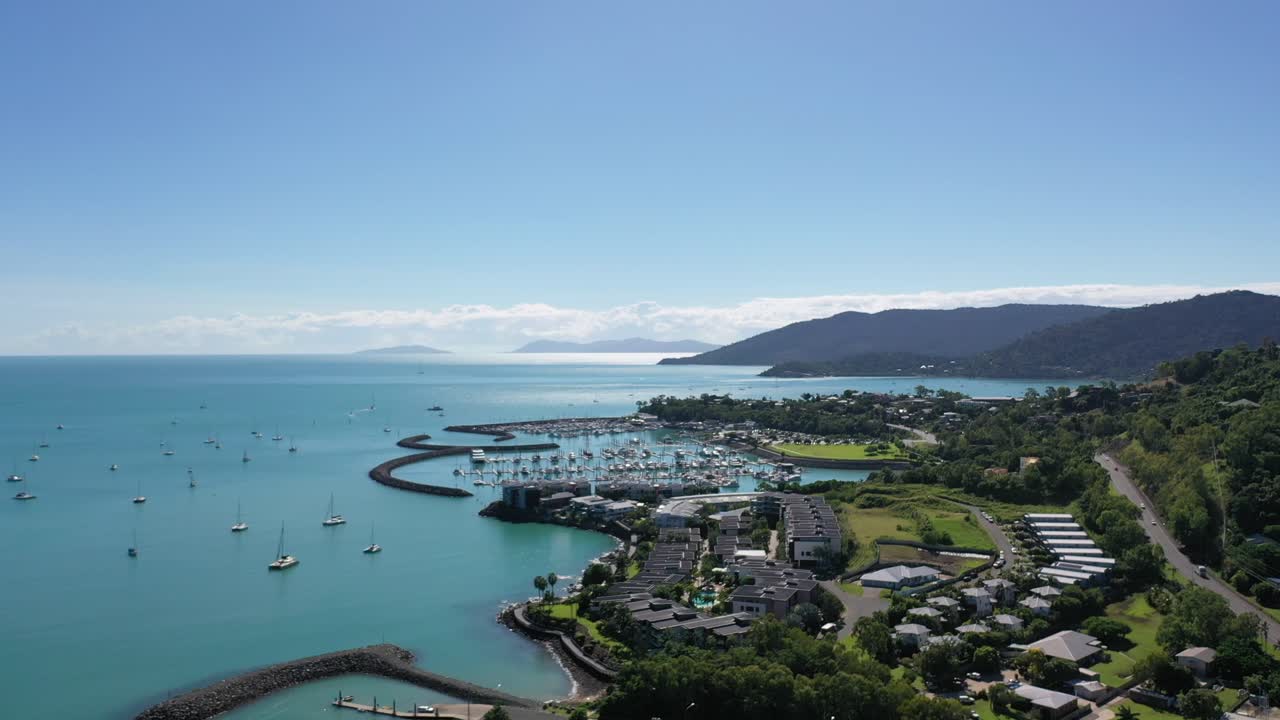 Aerial drone shot over Airlie Beach, Queensland - showing vast marina area with mountain range in background, yachts and boats on bright sunny day.