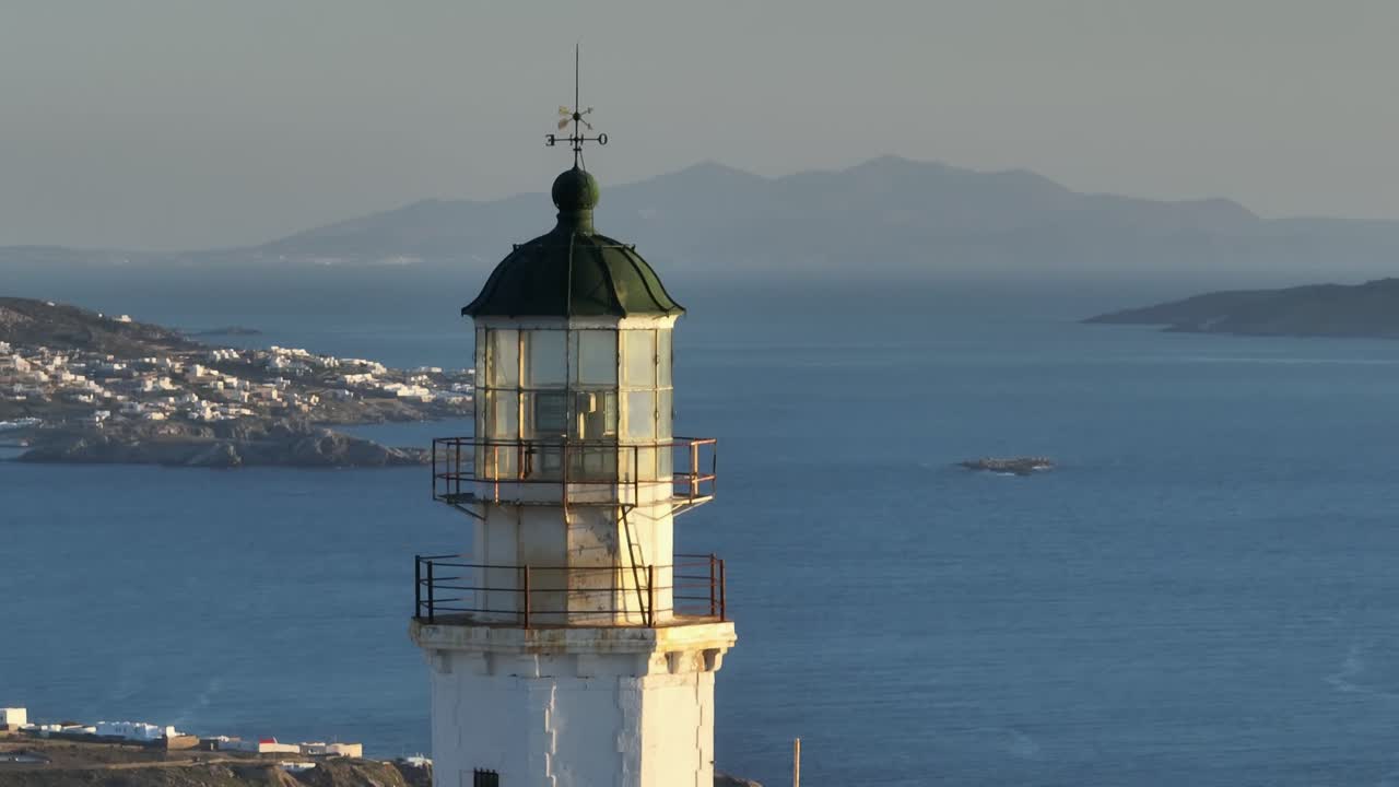 Drone orbits Armenistis Lighthouse using telephoto lens, revealing detailed view of Mykonos Chora in the background against the Aegean landscape