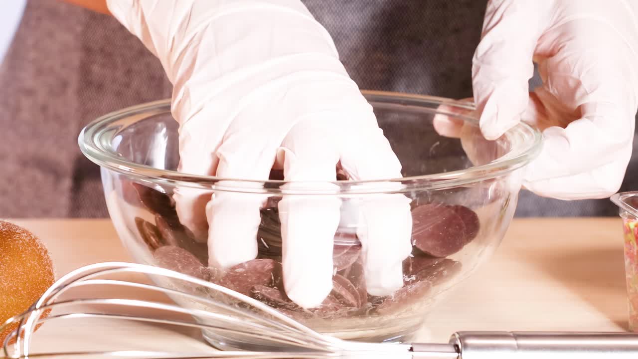 Close-up of gloved hands arranging chocolate discs in a glass bowl on a kitchen counter.