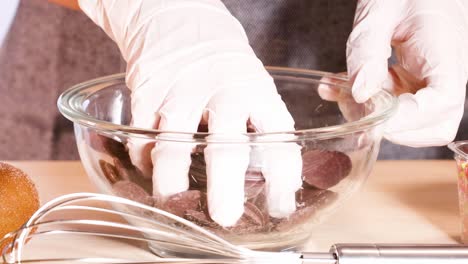close-up of gloved hands arranging chocolate discs in a glass bowl on a kitchen counter.