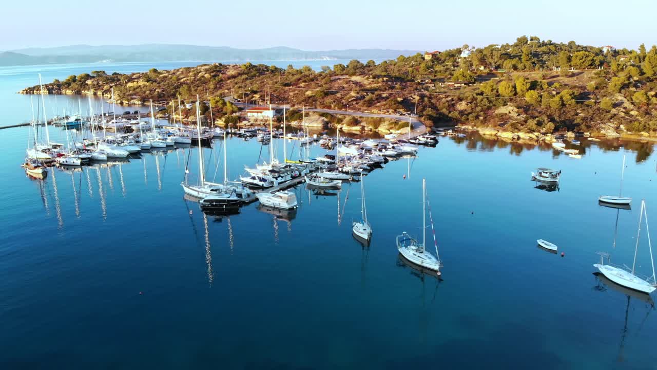 Aerial drone view of Aegean sea port with multiple moored yachts at piers, greenery, blue water, sunset, Greece