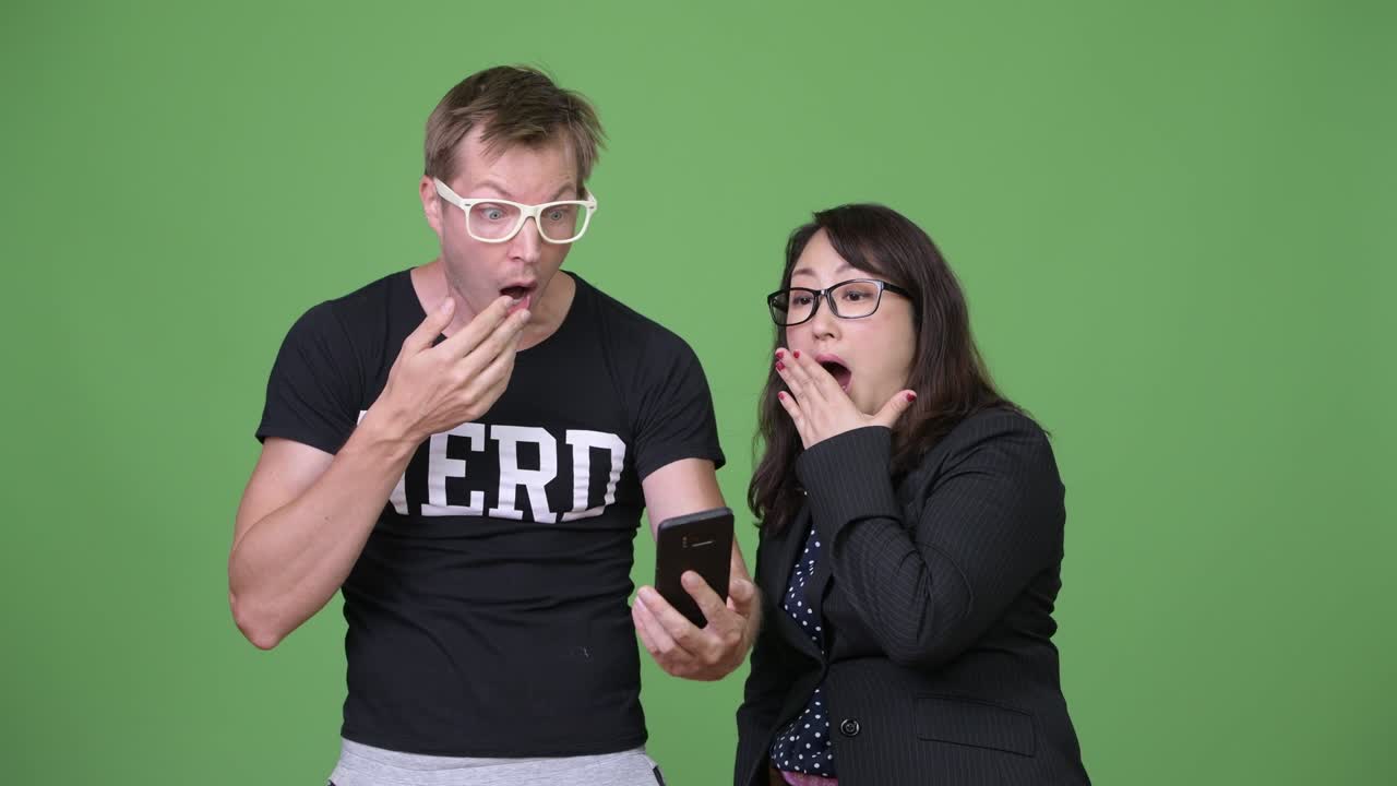 Mature Asian businesswoman and young Scandinavian nerd man using phone while looking shocked together