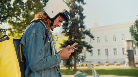 A-courier-guy-with-curly-hair-in-a-denim-shirt-walks-through-a-summer-park,-looks-around-and-looks-at-his-phone-for-navigation-and-gets-on-his-moped-with-a-large-yellow-bag-and-leaves-to-deliver-his-order-in-the-summer-city