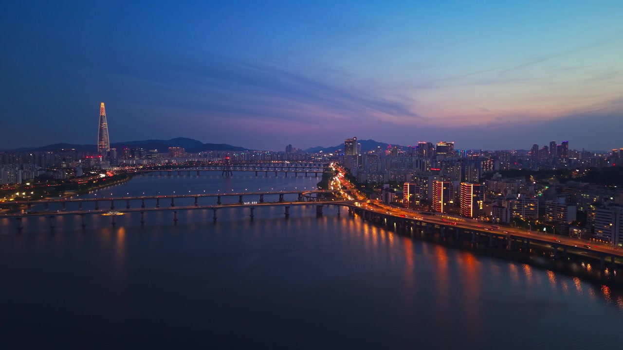 Ripresa aerea in allontanamento sul fiume Han all'ora blu, che rivela i ponti illuminati di Cheonho e Gwangjin, la scintillante superstrada Gangbyeonbuk-ro e la Lotte World Tower sullo skyline crepuscolare di Seul.
