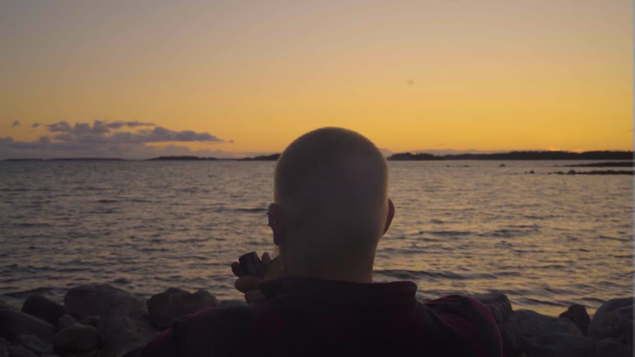 joven adulto sentado solo frente a las aguas mar océano río teniendo un sentido de pertenencia un fumar en el estilo anticuado una pipa llena de tabaco de calidad maravilloso lugar de puesta de sol