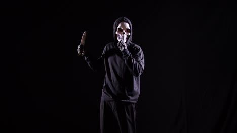 scary man in the hooded sweatshirt wearing halloween mask gesturing silence while holding a knife on the black background