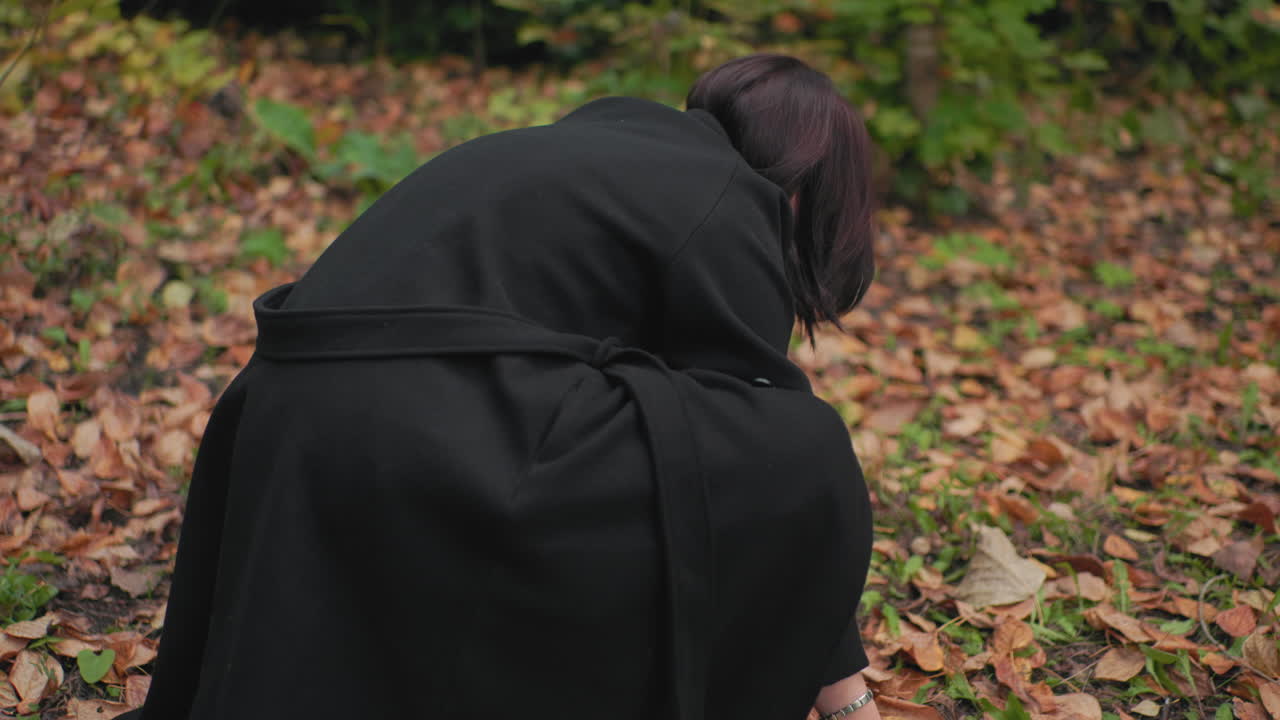 검은색 코트를 입은 숲 방문객이 낙엽으로 덮인 땅에 웅크려 앉아 조용한 가을 숲 속 산책을 하는 동안 두 손으로 마른 잎을 모아 더미를 만들고 있어요. 부드러운 햇빛 아래 흙색이 감도는 조용하고 깔끔한 순간이에요.