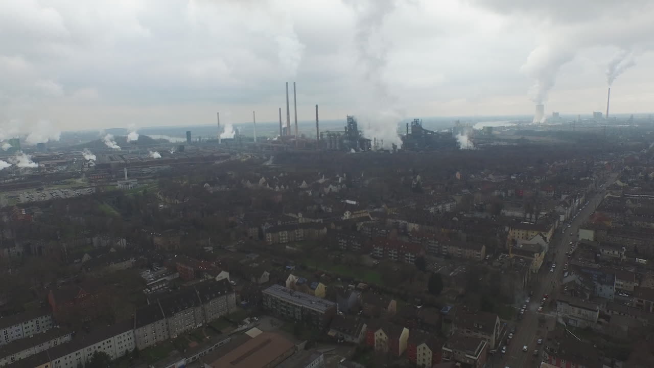 Aerial backward shot over an industrial German city with multiple smoking chimneys under heavy clouds and a dark urban atmosphere