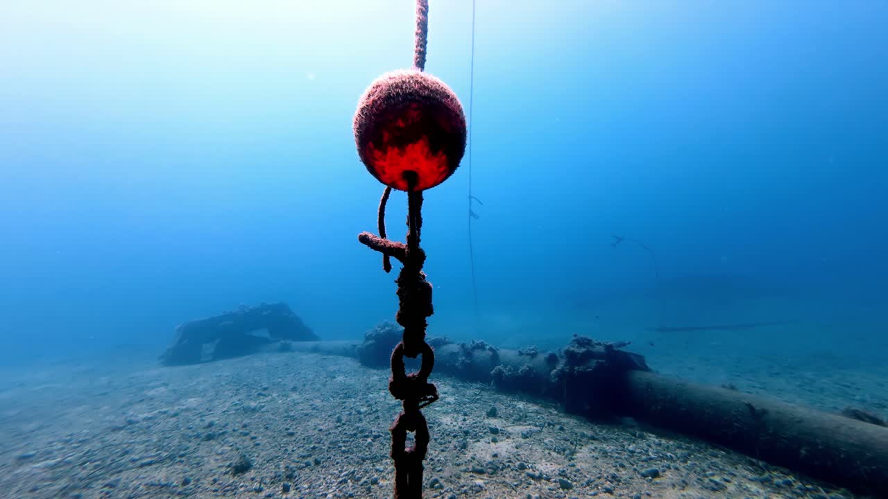 晴れた夏のマルタ島ゴゾ沖、地中海の砂地の海底に、赤いブイと工業用パイプが付属した沈んだ錨の鎖が映し出されている。透明な青い水とローアングルからの眺めが特徴。