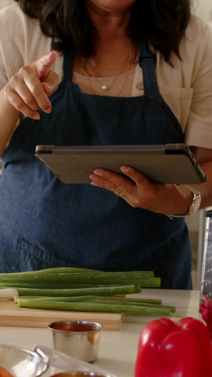 Video vertical: Mujer con delantal abriendo una receta en la tablet, tocándola y colocando cebolletas en la encimera.