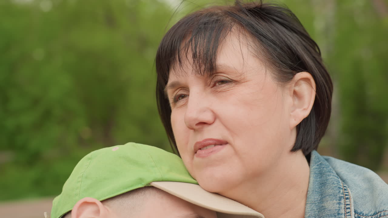Woman Resting Head On Young ManS Shoulder, CloseUp Portrait Of Caregiver In Denim Jacket Holding Green Cap Child In Park, Eyes Closed, Wind Brushing Hair, Conveying Quiet Fatigue, Comfort And Tender