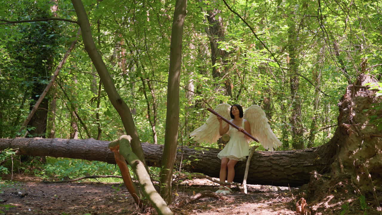 Angel woman with white wings sits on fallen tree holding wooden staff in sunlit forest, surrounded by bright green leaves, atmosphere calm and magical