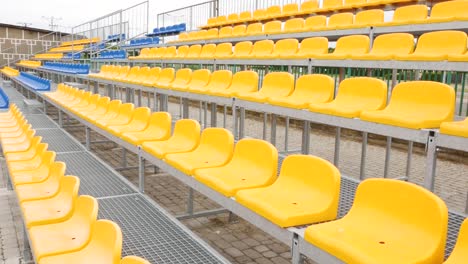 empty vacant stadium stands, chairs, plastic yellow arena spectator seats on a small football soccer field, daytime, pan, nobody. no people, sports event site, fans absence, rows of seats outdoors