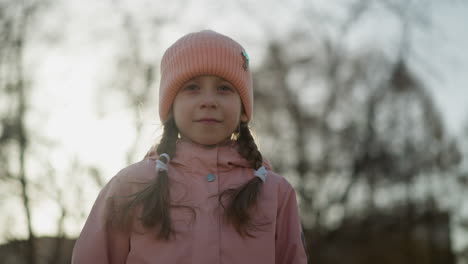 front view of a little girl wearing a pink cap and pink jacket, walking and smiling. her braided hair and the warm sunlight create a charming and joyful atmosphere as she enjoys her day outdoors