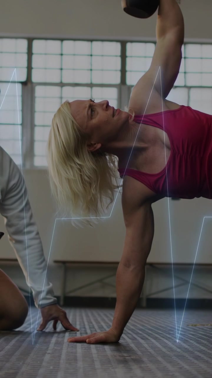 Vertikales Video: Trainer gibt Anweisungen an Frau im pinken Tanktop, die im Fitnessstudio eine Kettlebell rudernd hochhebt, mit EKG-Linie für die Rumpfmuskulatur.