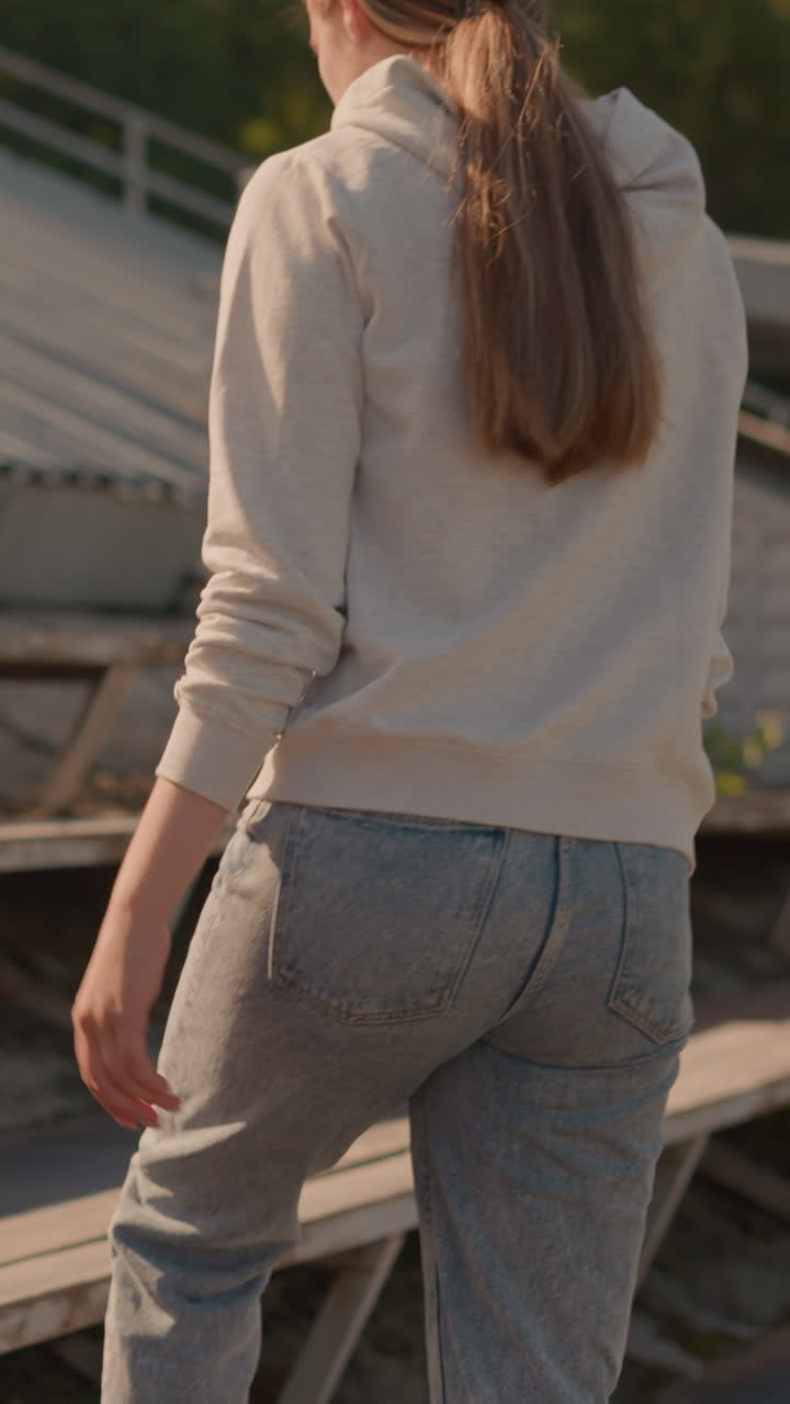 Back view of young woman in casual hoodie and jeans walking towards bleachers in outdoor stadium, soft sunlight highlights her relaxed posture as she approaches rustic seating area