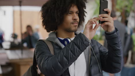 Porträt-Eines-Jungen-Studenten-Gemischter-Abstammung,-Der-Mit-Smartphone-Kameratechnologie-Vor-Einem-Lebendigen-Stadthintergrund-Fotografiert