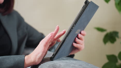Businesswoman in grey suit using digital tablet on couch with hand gesture mid-air, possibly interacting with screen or video calling, indoors in relaxed work-from-home or casual business setting