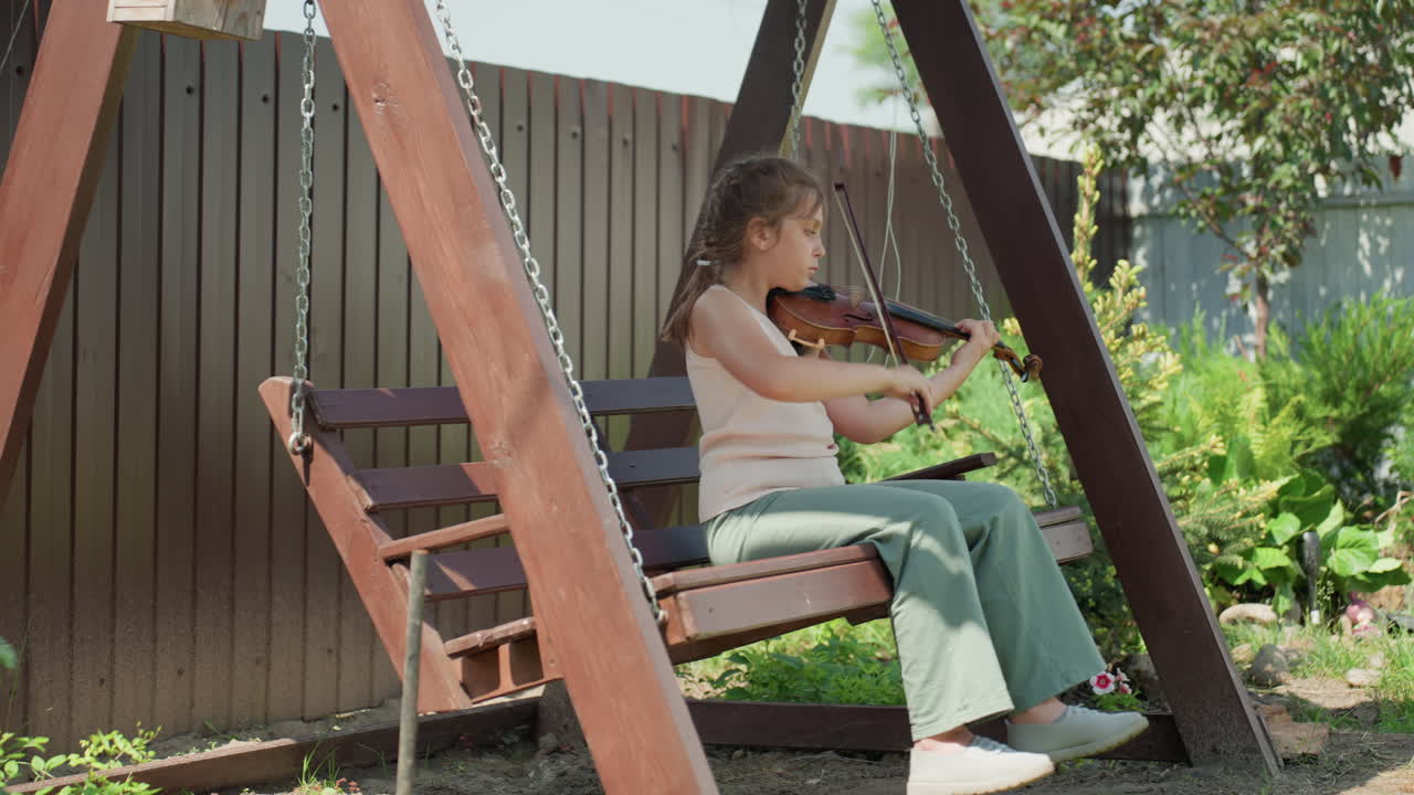 Young Woman Playing Violin Outside, Young Girl Rehearsing With Violin On Wooden Swing Amidst Green Foliage, Female Violinist Enjoying Peaceful Outdoor Practice Session In Leafy Garden Environment