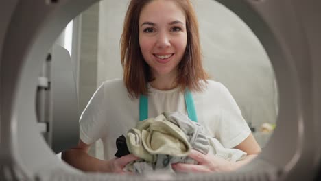 view-from-inside-the-washing-machine-portrait-of-a-brunette-girl-in-a-white-T-shirt-who-loads-dirty-things-into-the-washing-machine-darkening-the-light-with-these-clothes-in-a-modern-apartment