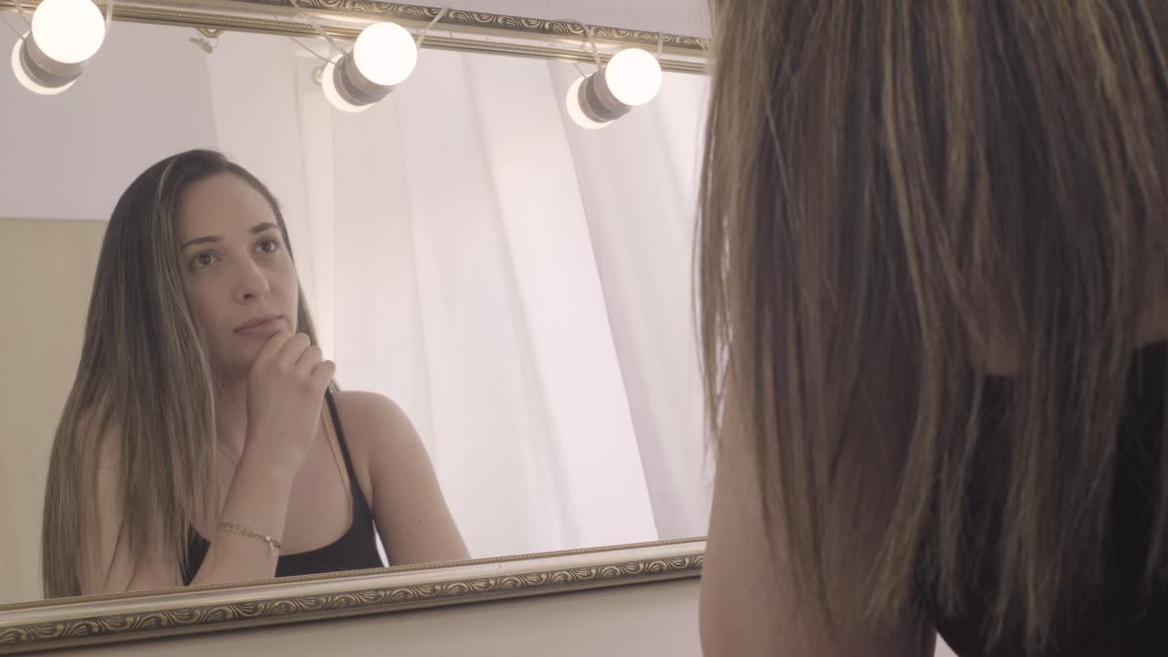 Giovane ragazza si guarda allo specchio con luci per il trucco, pensierosa.