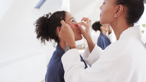 happy biracial lesbian couple in bathrobes applying under eye masks in sunny bathroom, slow motion