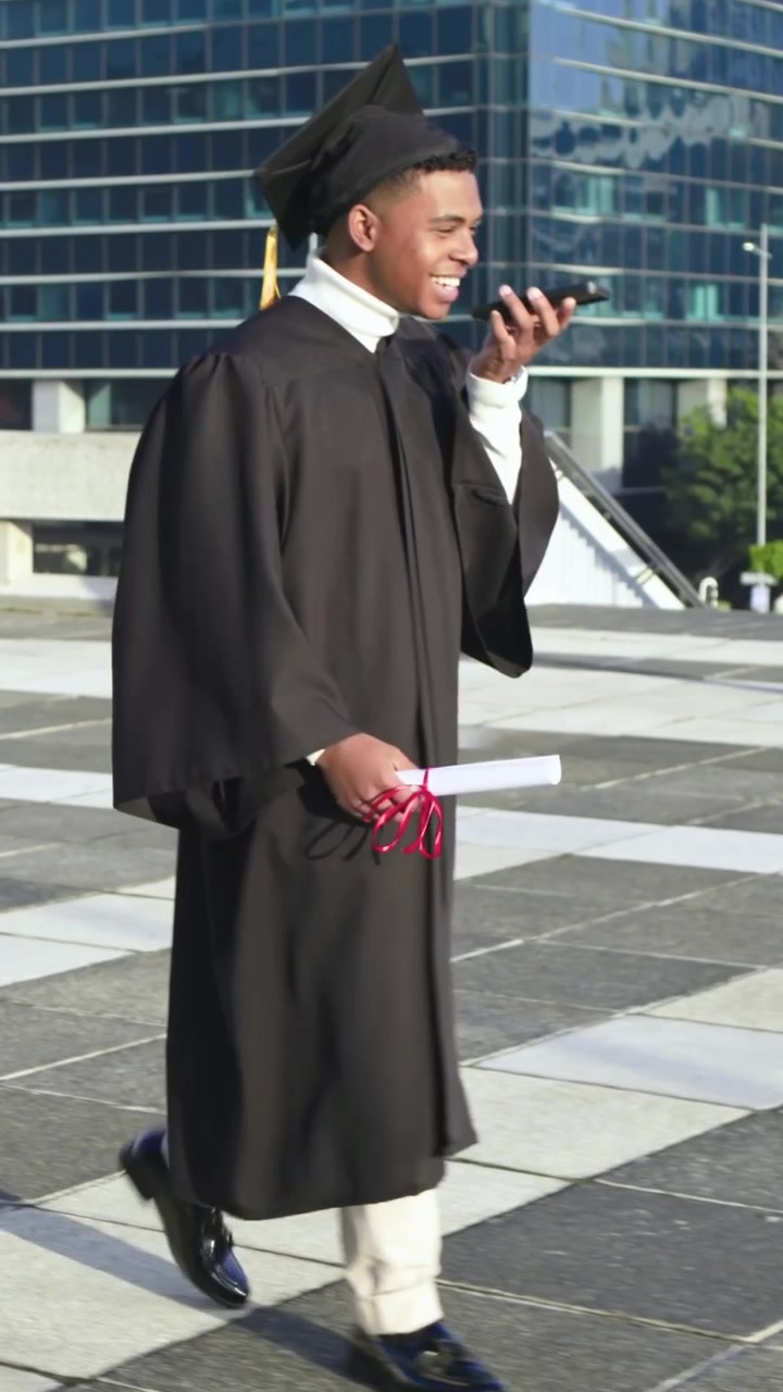 Vertical video: Reacting to phone ping grad in gown, cap walking on rooftop with diploma and phone