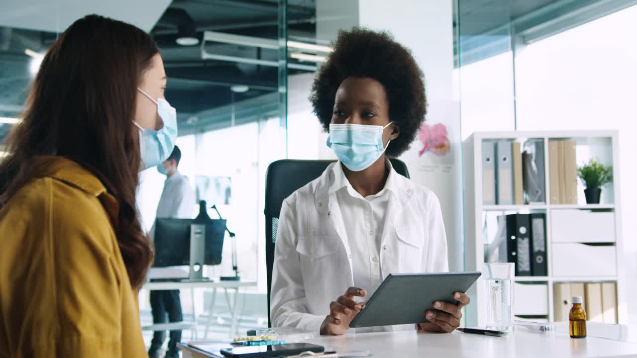 Close-up view of African American female doctor in medical mask holding a tablet and explaining to female patient treatment for coronavirus in medical consultation