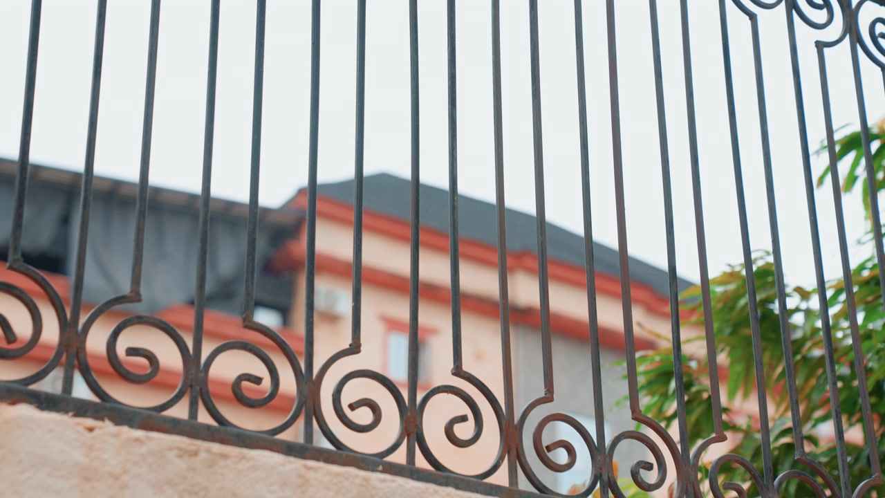 Cancello e ringhiera in ferro battuto ornati davanti a una facciata residenziale, dettagli in metallo a volute con scorci di tetto e vegetazione, luce diurna soffusa, atmosfera domestica intima che suggerisce privacy e un ingresso sicuro.