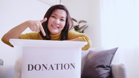 Frau,-Die-Auf-Eine-Spendenbox-Zeigt-Und-Menschen-Einlädt,-Sich-Der-Kampagne-Anzuschließen,-Um-Unbenutzte-Gegenstände-Zu-Spenden